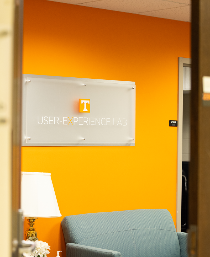 An orange wall with a sign on it for the User Experience Lab with a light blue couch beneath 