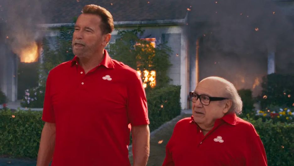 A still of the Super Bowl State Farm commercial shows Arnold Schwarzenegger on the left and Danny DeVito on the right, both wearing matching red State Farm branded polos, while a house in the background is visibly on fire.