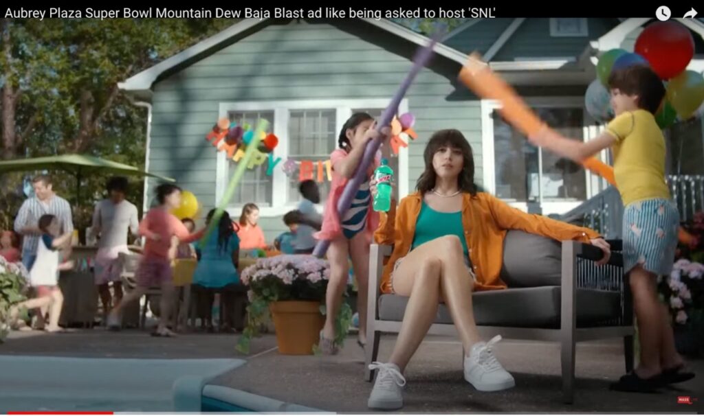 A screenshot of a commercial shows a bored-looking Aubrey Plaza sitting on a bench while children play with pool noodles all around her and people in the background celebrate at a birthday party.