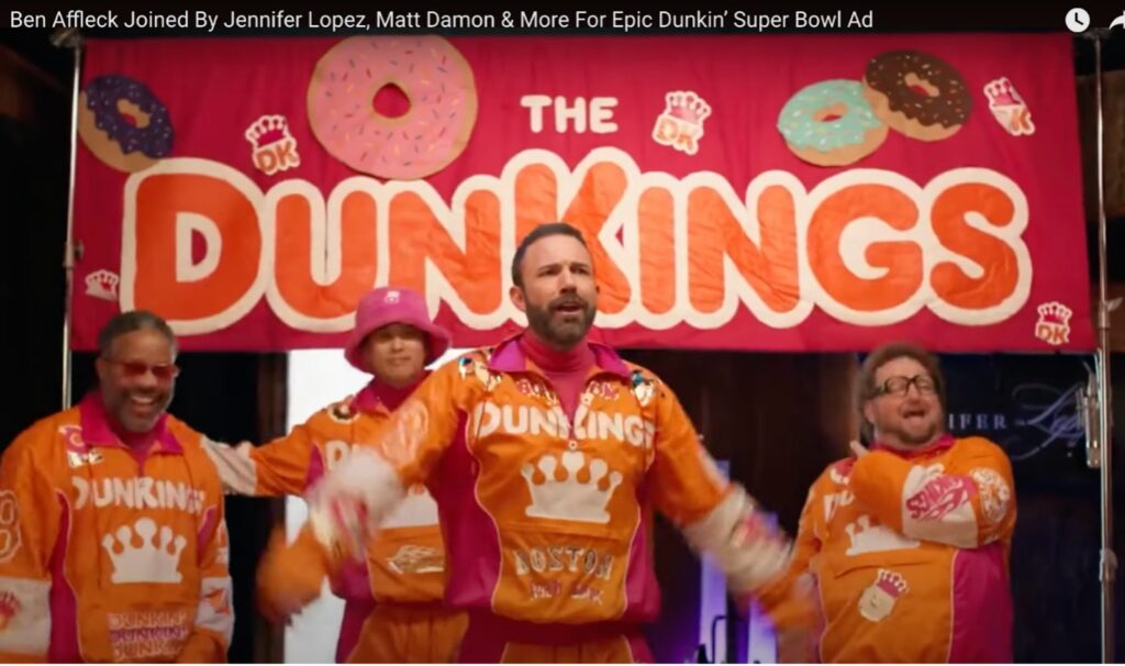 A still of a Dunkin' Donuts commercial shows Ben Affleck, front and center, with three other men in the background all dressed in matching orange and pink Dunkin' Donuts branded track suits with a big banner stating "The DunKings" hanging above them in the background.